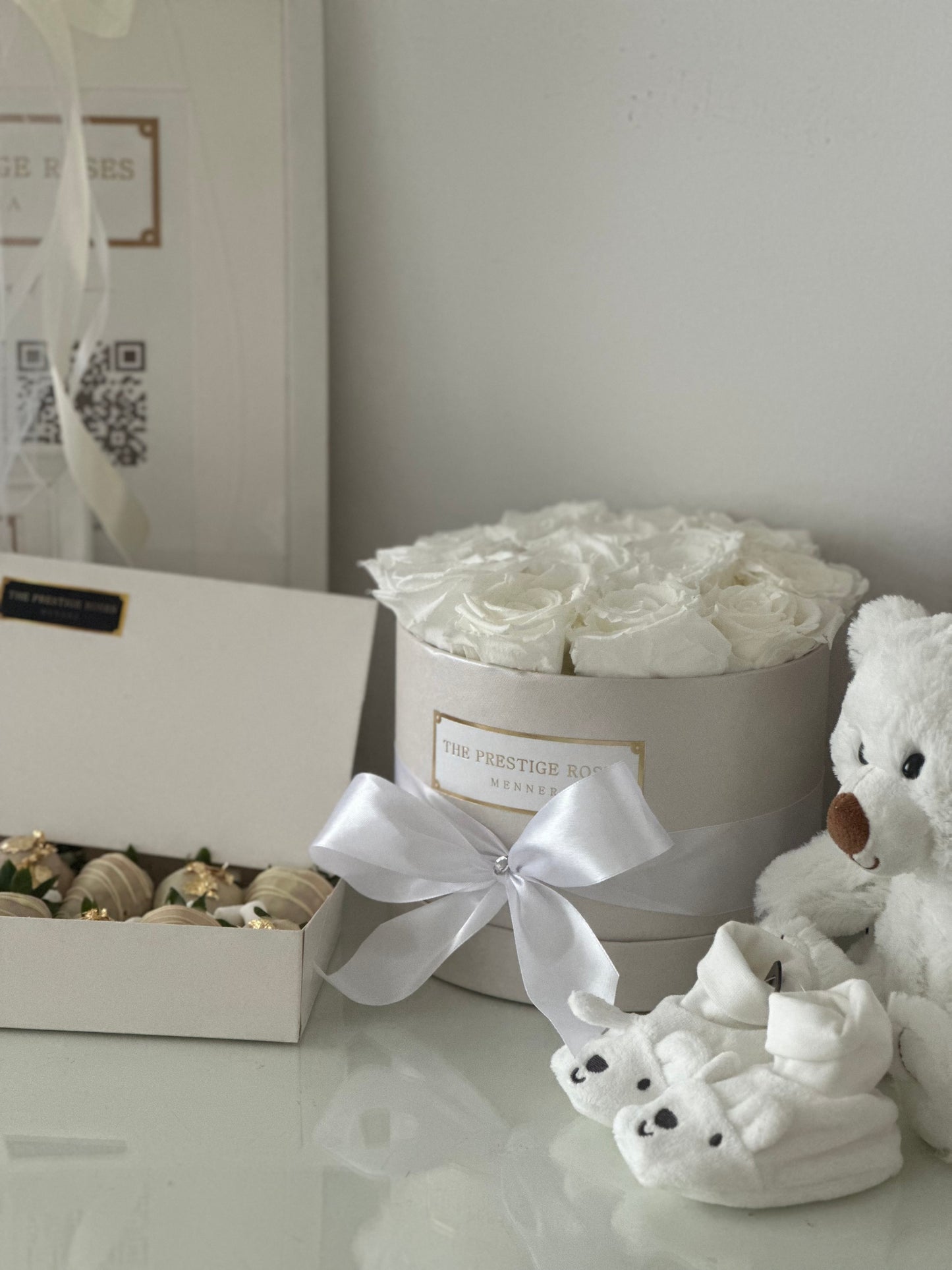 Caja cilíndrica blanca con flores preservadas y accesorios de bebé como parte de un set de regalo exclusivo.