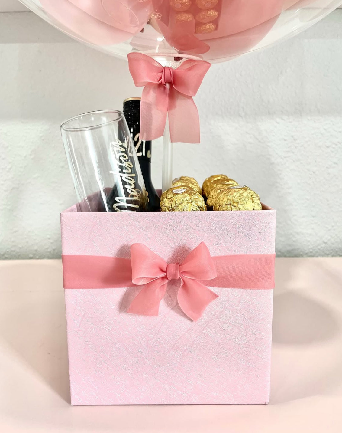 Detalle de la caja rosa del set de regalo con lazo satinado, chocolates Ferrero Rocher, copa personalizada y botella mini de vino espumoso – ideal para cumpleaños elegantes de The Prestige Roses Madrid.
Close-up of the pink gift box with satin bow, Ferrero Rocher chocolates, personalized glass and mini sparkling wine bottle – perfect for elegant birthdays by The Prestige Roses Madrid.