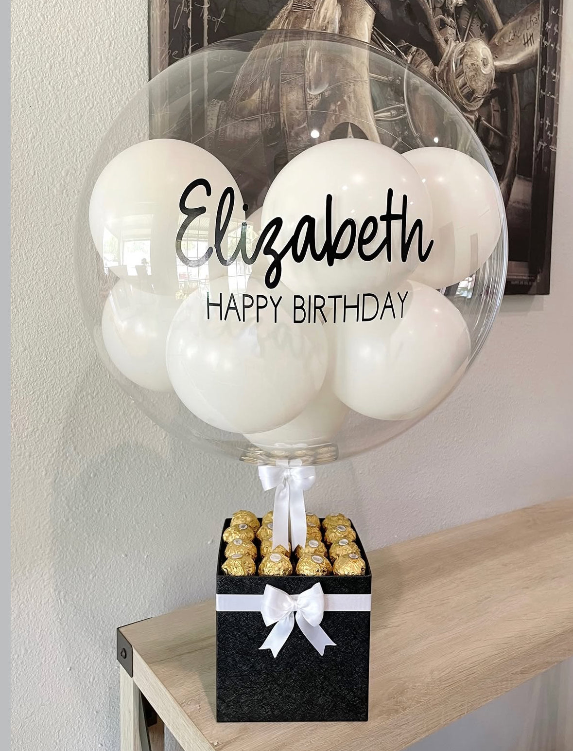 Set de regalo negro con globo transparente personalizado “Happy Birthday Elizabeth”, decorado con mini globos blancos, lazo satinado blanco y chocolates Ferrero Rocher – un detalle elegante y sofisticado de The Prestige Roses Madrid.
Black gift box with personalized “Happy Birthday Elizabeth” transparent balloon, white mini balloons, satin bow and Ferrero Rocher chocolates – a refined and elegant gift by The Prestige Roses Madrid.