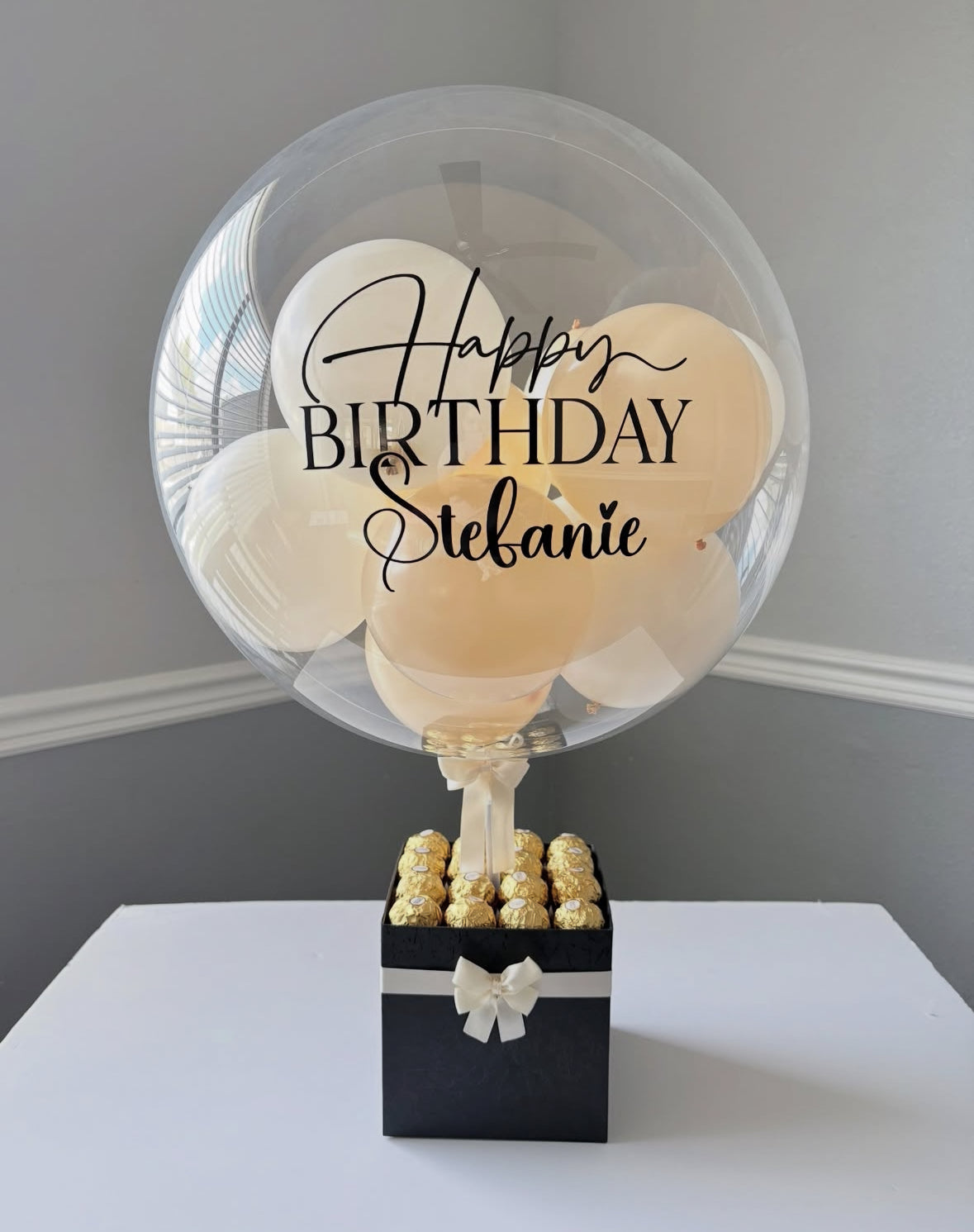 Set de regalo negro con globo transparente personalizado y mini globos dorados, decorado con lazo blanco y chocolates Ferrero Rocher – un detalle elegante de The Prestige Roses Madrid.
Black and gold personalized balloon gift box with white bow and Ferrero Rocher chocolates – a luxury and elegant detail by The Prestige Roses Madrid.