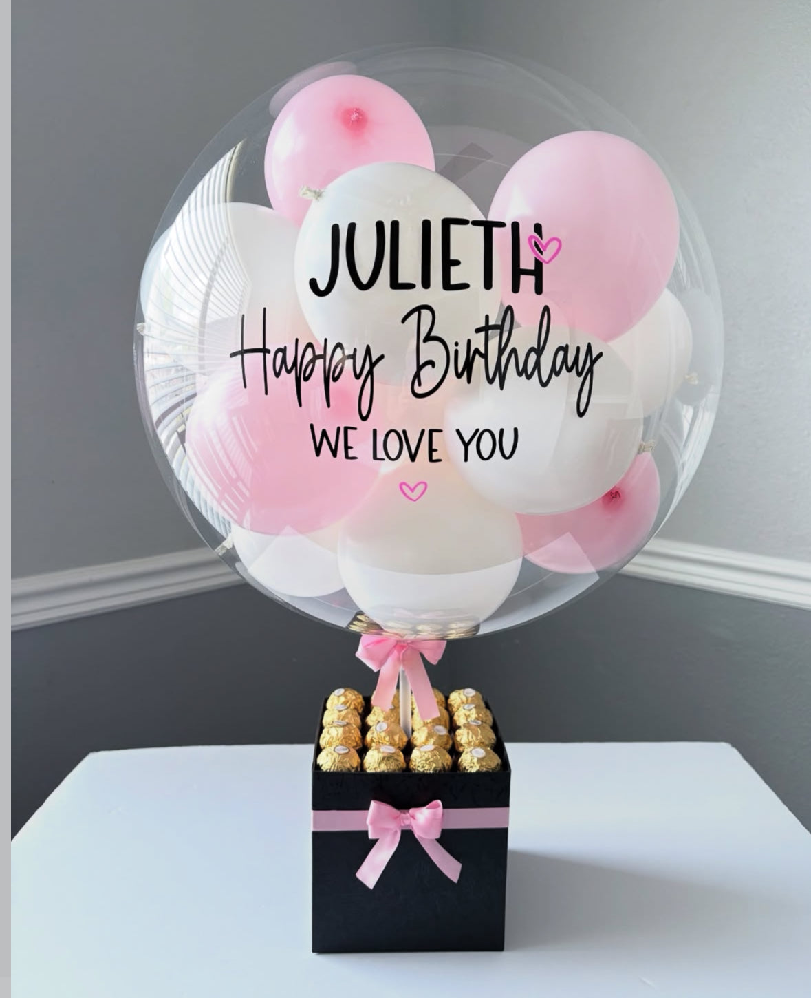 Set de regalo negro con globo transparente personalizado “Happy Birthday Julieth”, decorado con mini globos rosa y blanco, lazo satinado rosa y chocolates Ferrero Rocher – detalle elegante y moderno de The Prestige Roses Madrid.
Black gift box with personalized “Happy Birthday Julieth” transparent balloon, pink and white mini balloons, satin bow and Ferrero Rocher chocolates – a modern and elegant luxury gift by The Prestige Roses Madrid.