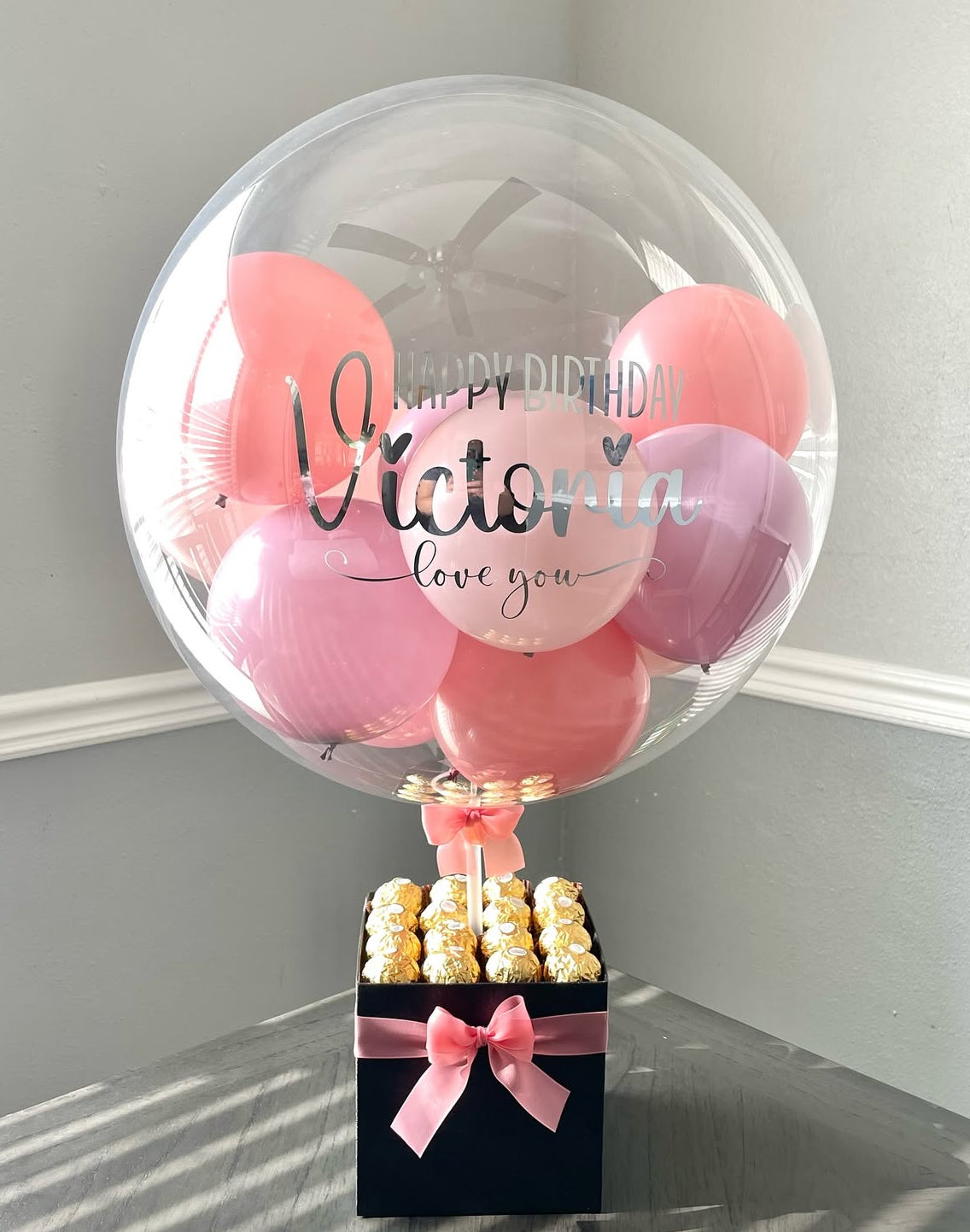 Set de regalo negro con globo transparente personalizado “Happy Birthday Victoria – Love You”, decorado con globos en tonos rosa y fucsia, lazo satinado rosa y chocolates Ferrero Rocher – un detalle romántico y sofisticado de The Prestige Roses Madrid.
Black gift box with personalized “Happy Birthday Victoria – Love You” transparent balloon, pink and fuchsia mini balloons, satin pink bow and Ferrero Rocher chocolates – a romantic and sophisticated gift by The Prestige Roses Madrid.