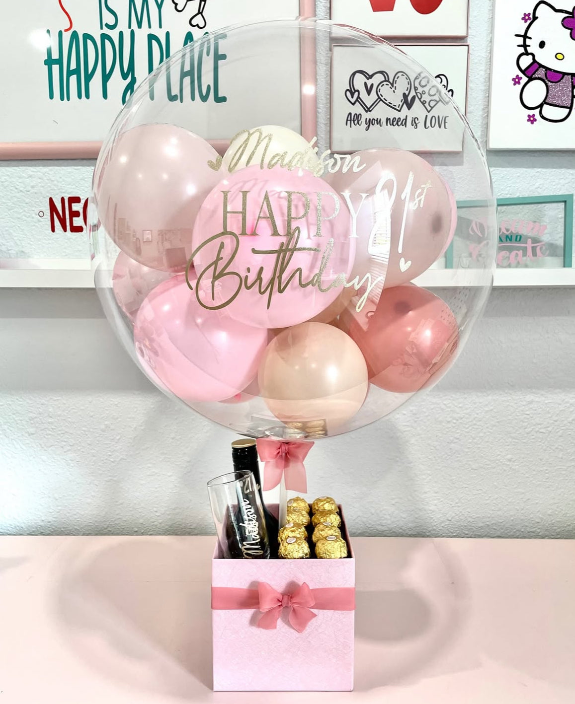 Set de regalo rosa con globo transparente personalizado “Happy 21st Birthday Madison”, decorado con globos en tonos rosa y champagne, botella mini de vino espumoso, copa personalizada y chocolates Ferrero Rocher – un regalo de cumpleaños elegante y festivo de The Prestige Roses Madrid.
Pink gift box with personalized “Happy 21st Birthday Madison” transparent balloon, pink and champagne mini balloons, mini sparkling wine bottle, personalized glass and Ferrero Rocher chocolates – a festive and elegant birthda