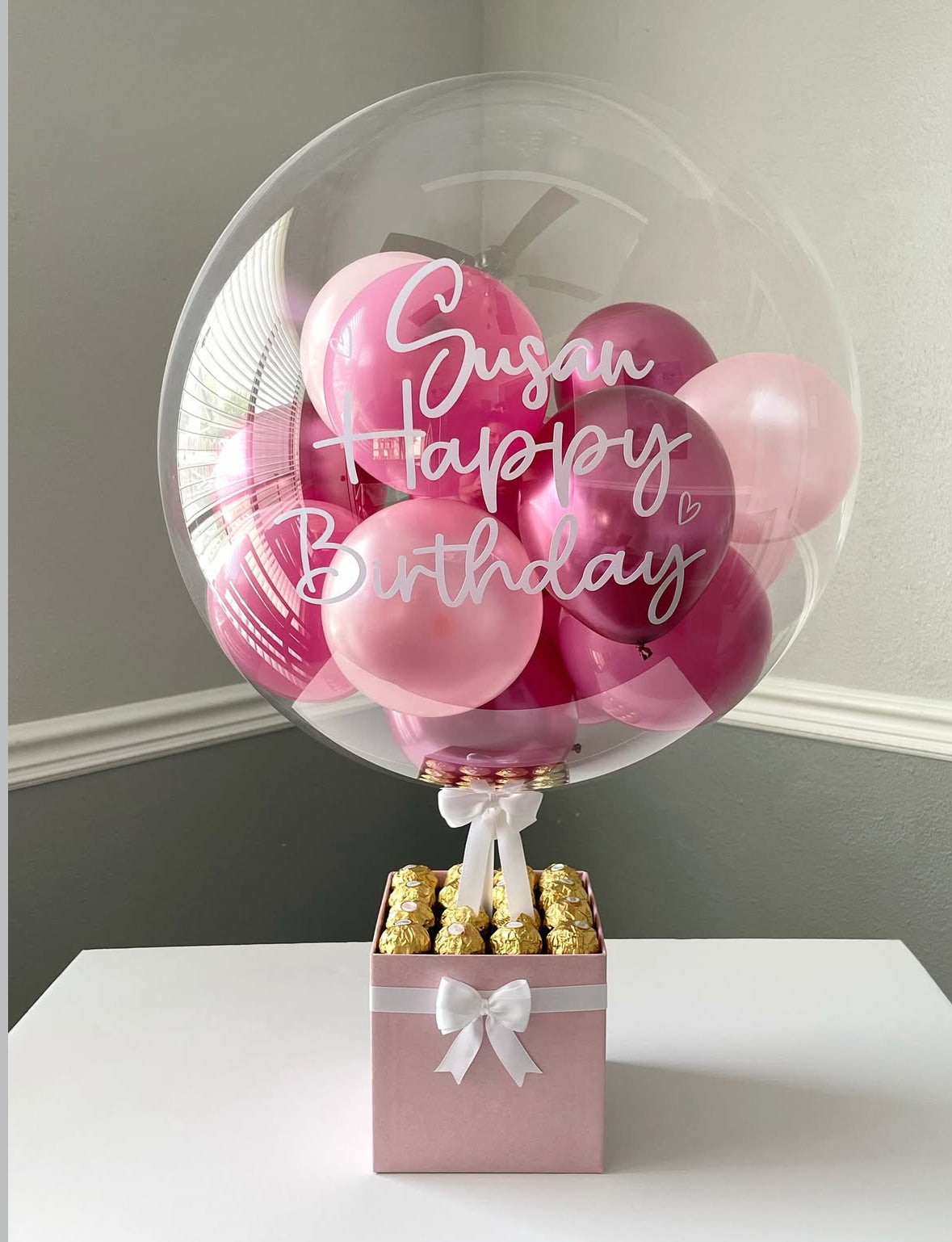 Set de regalo rosa con globo transparente personalizado “Happy Birthday Susan”, decorado con globos en tonos rosa fucsia y pastel, lazo blanco y chocolates Ferrero Rocher – un detalle dulce y elegante de The Prestige Roses Madrid.
Pink gift box with personalized “Happy Birthday Susan” transparent balloon, fuchsia and pastel pink mini balloons, white bow and Ferrero Rocher chocolates – a sweet and elegant birthday detail by The Prestige Roses Madrid.