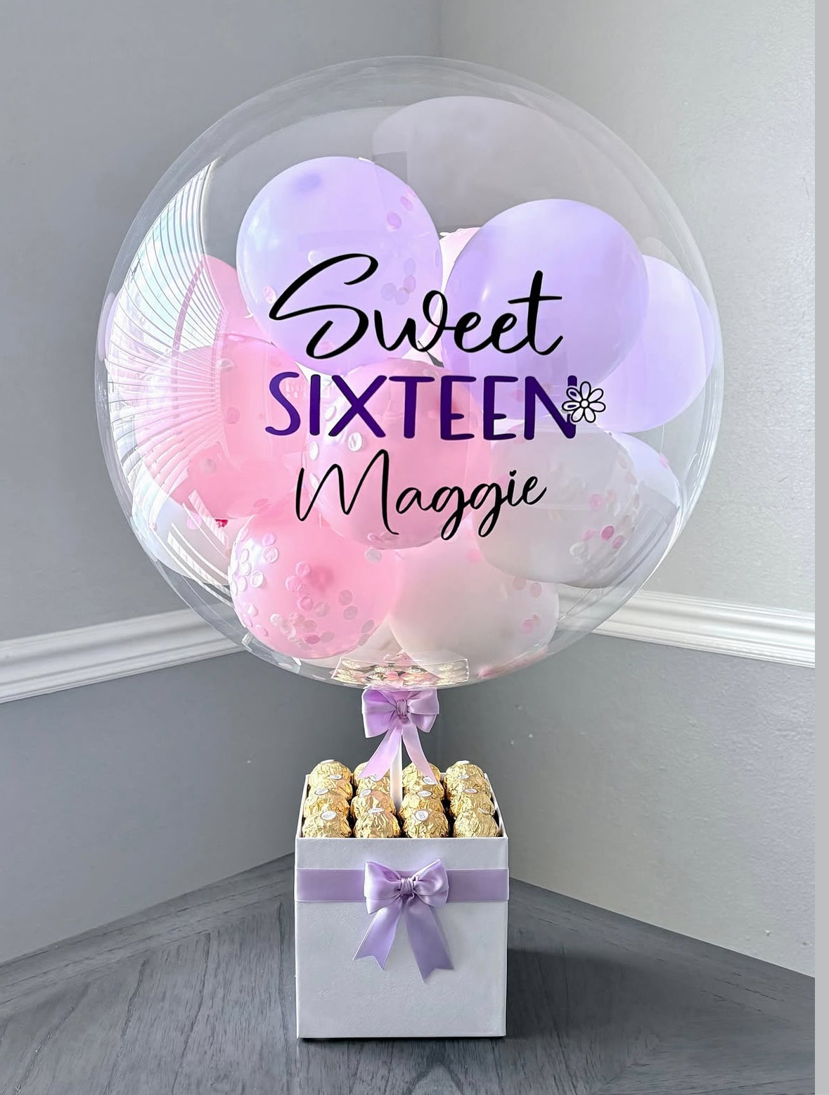Set de regalo blanco con globo transparente personalizado “Sweet Sixteen” en tonos rosa y lila, decorado con lazo satinado y chocolates Ferrero Rocher – detalle elegante y juvenil de The Prestige Roses Madrid.
White gift box with personalized “Sweet Sixteen” balloon in pink and lilac tones, satin bow and Ferrero Rocher chocolates – an elegant and youthful detail by The Prestige Roses Madrid.