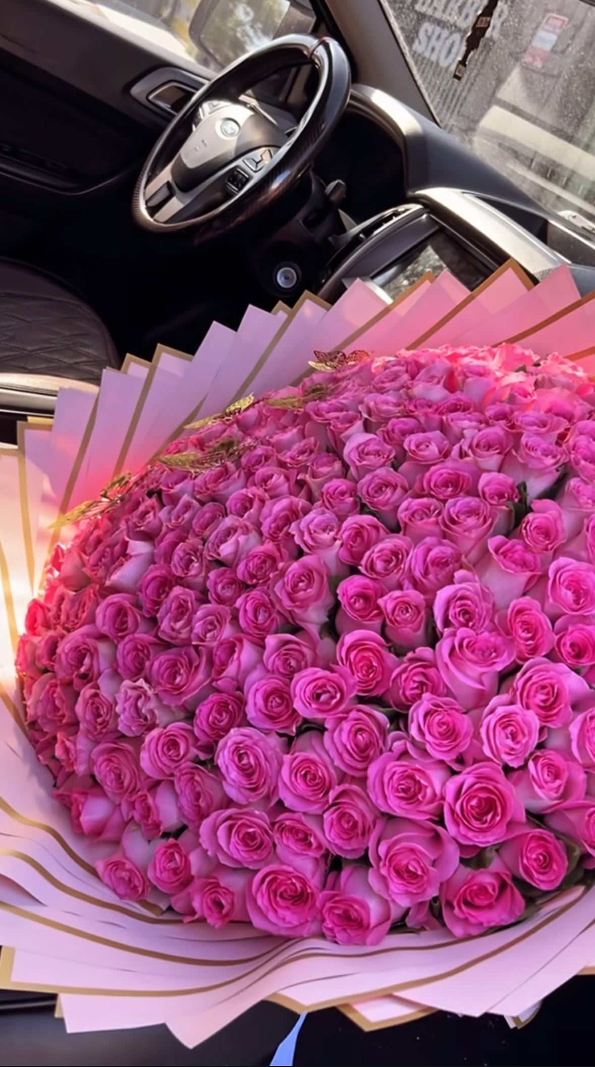 Ramo gigante con 200 rosas fucsia, envuelto en papel rosa con borde dorado y lazo blanco, apoyado en el asiento del coche visto desde arriba.
Giant bouquet with 200 fuchsia roses, wrapped in pink paper with golden edges and white ribbon, placed on the car seat viewed from above.
