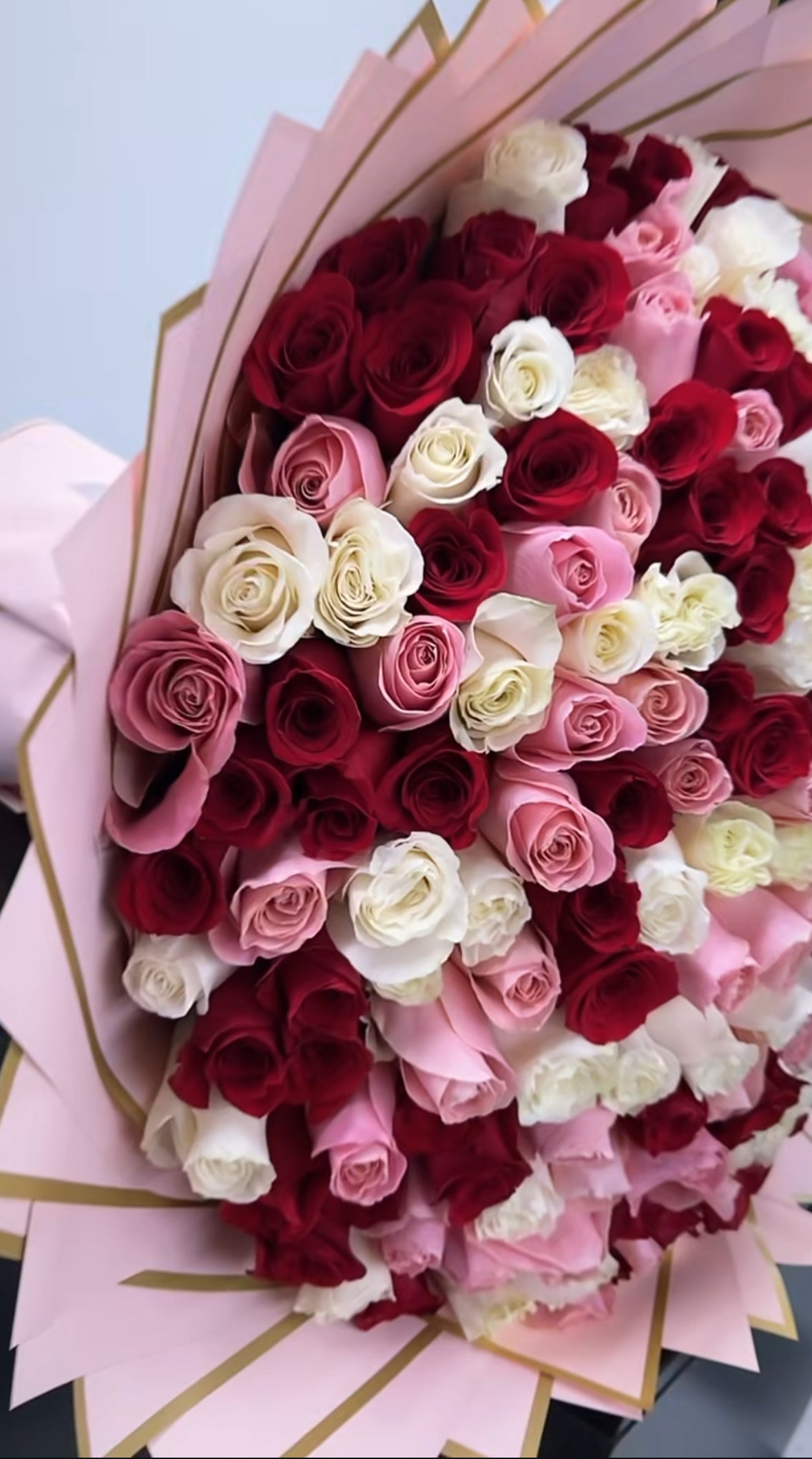 Ramo gigante de rosas rojas, rosas y blancas en forma redonda, envuelto en capas de papel rosa con borde dorado, estilo lujo.
Giant round bouquet of red, pink and white roses wrapped in layered pink paper with gold edges, luxury style.