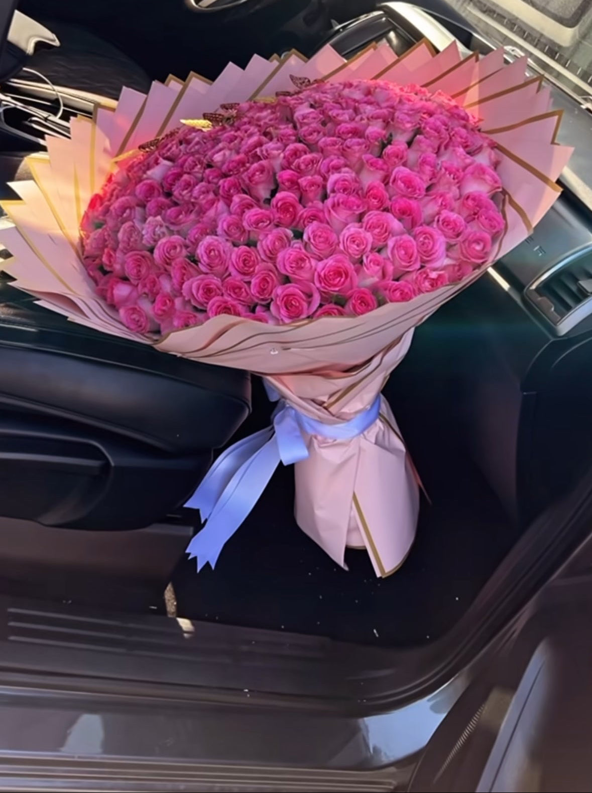 Ramo extragrande con 200 rosas rosa fucsia, envuelto en papel en capas con borde dorado y lazo blanco, colocado sobre el asiento de un coche.
 Extra-large bouquet with 200 fuchsia pink roses, wrapped in layered paper with golden edges and white ribbon, placed on a car seat.