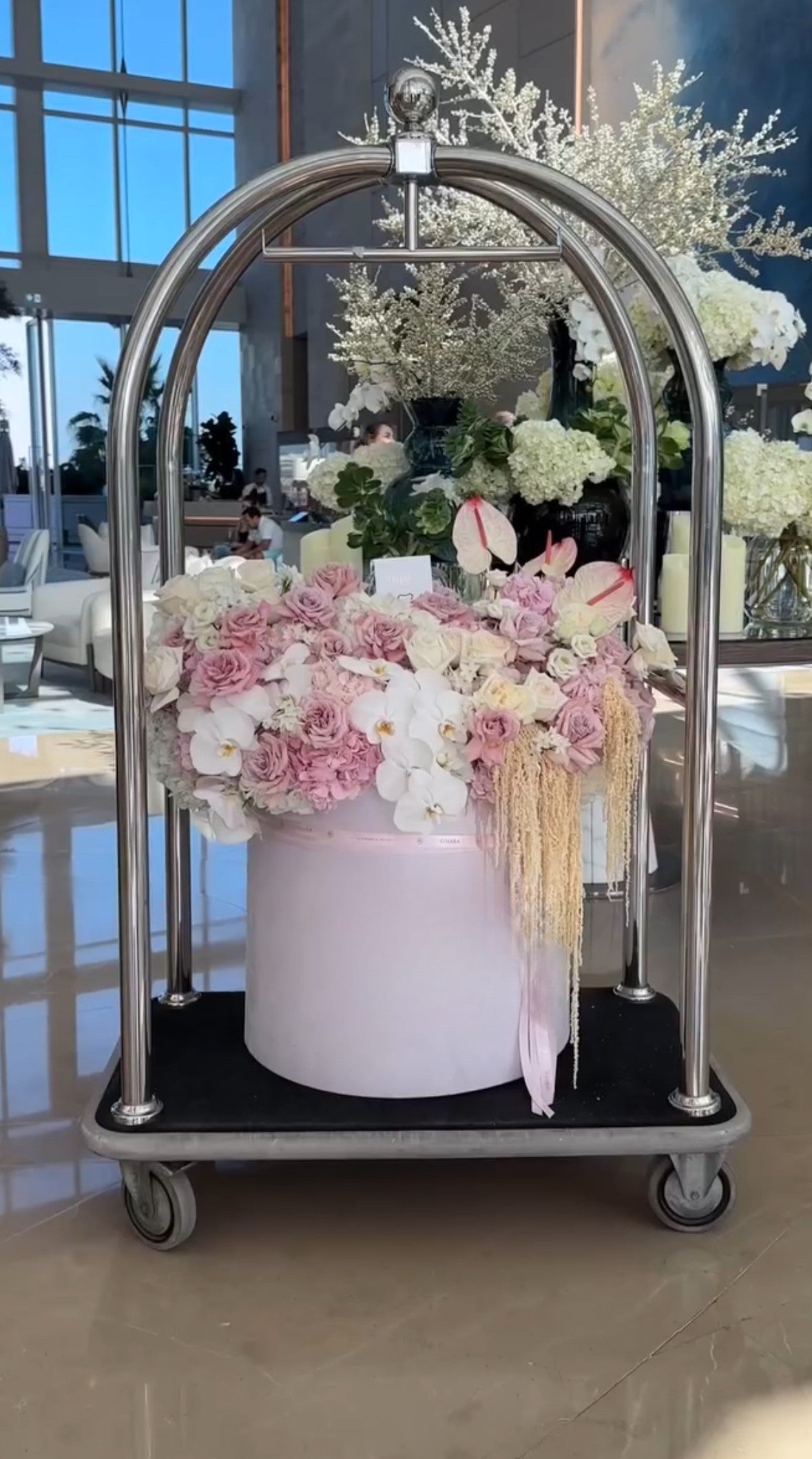 Caja de flores de lujo en tonos pastel sobre carrito de hotel en un lobby elegante con luz natural.