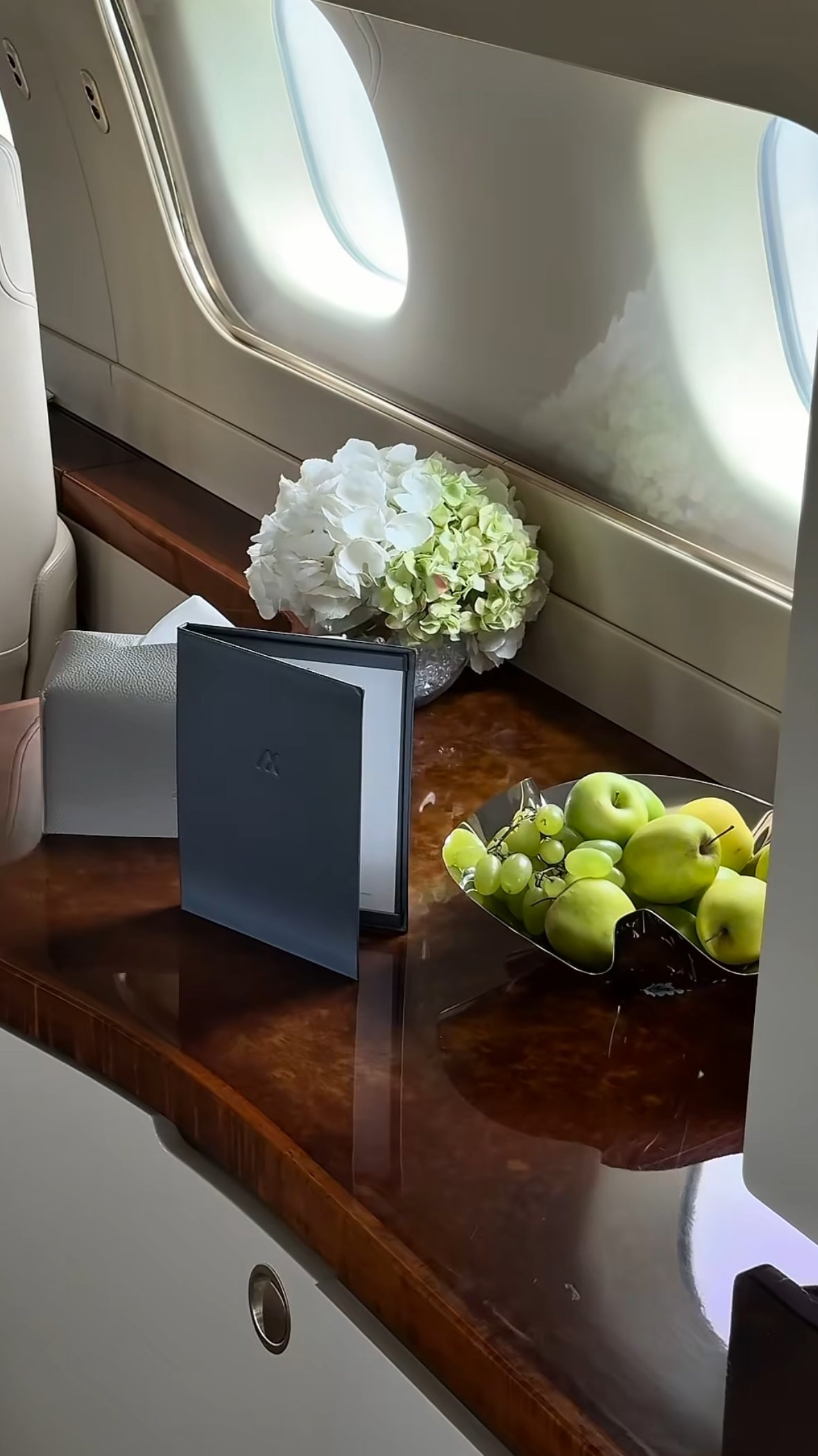 Detalle elegante junto a la ventana de un jet privado con hortensias blancas, manzanas verdes y caja de regalo de lujo.