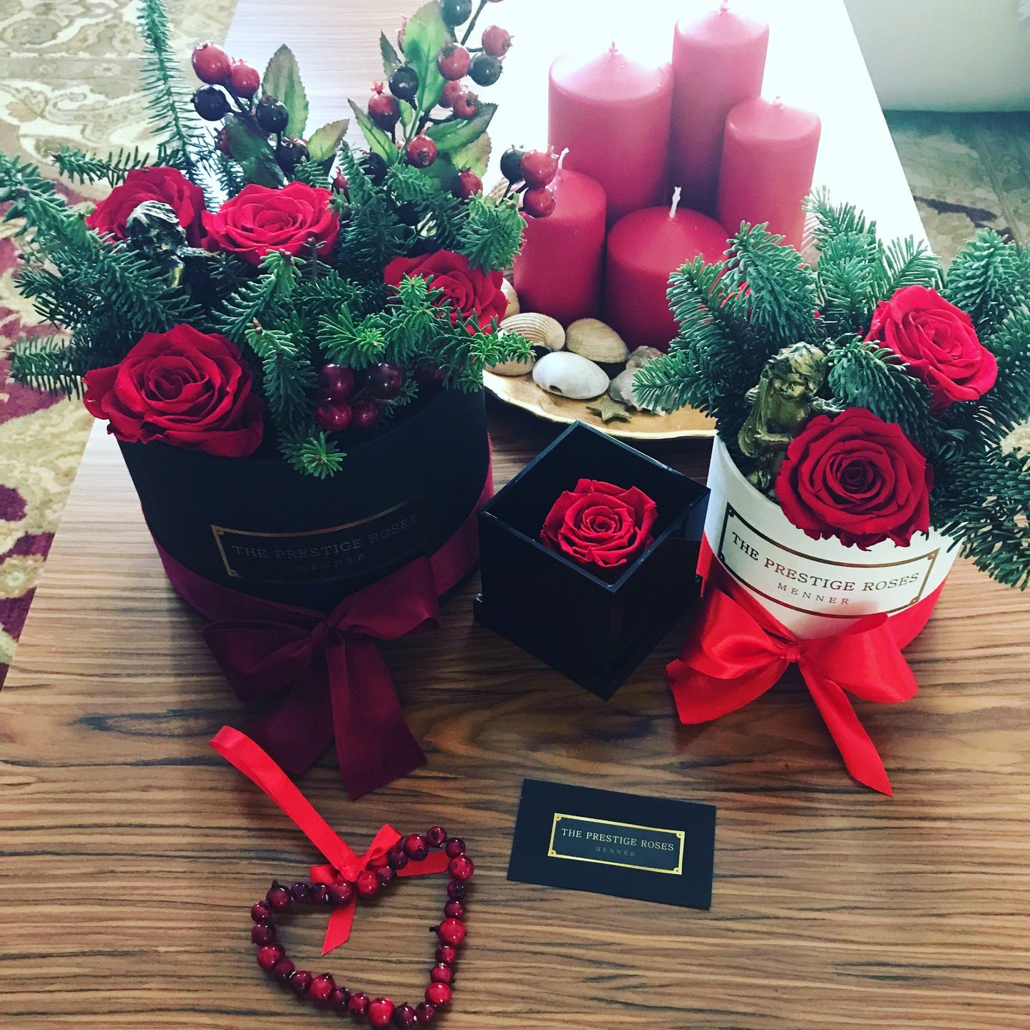  Arreglo navideño con cajas negras y blancas de rosas rojas preservadas, adornadas con ramas de pino, lazos rojos y detalles festivos.